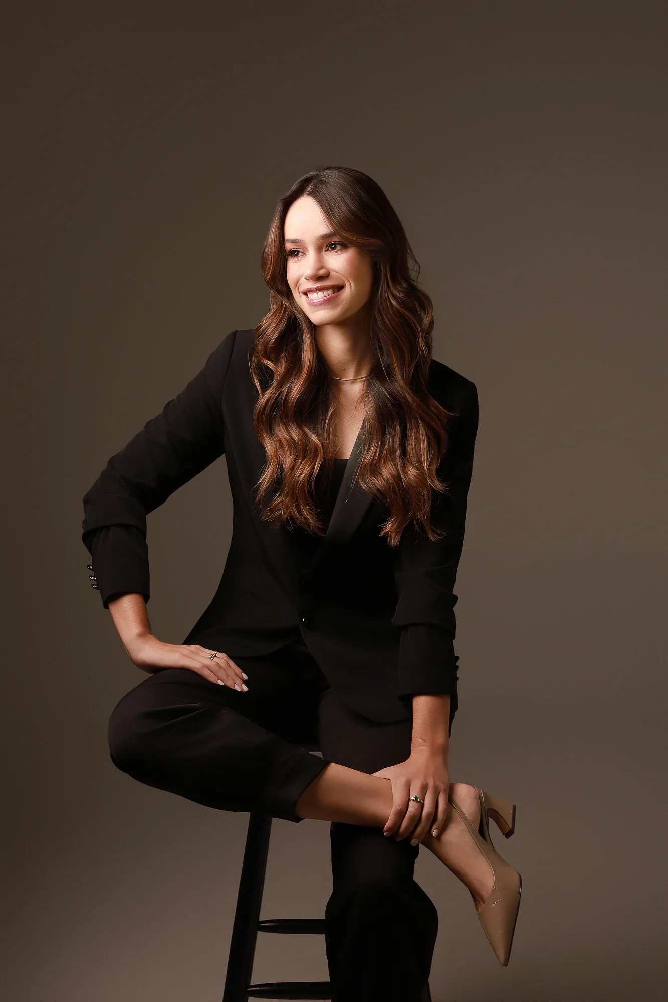 Dra. Mariana Sathler sentada em um banco, usando terno preto, diante de fundo marrom.